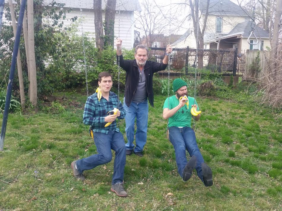 John and Jesse sit on a swingset eating bananas while Dave stands behind them.